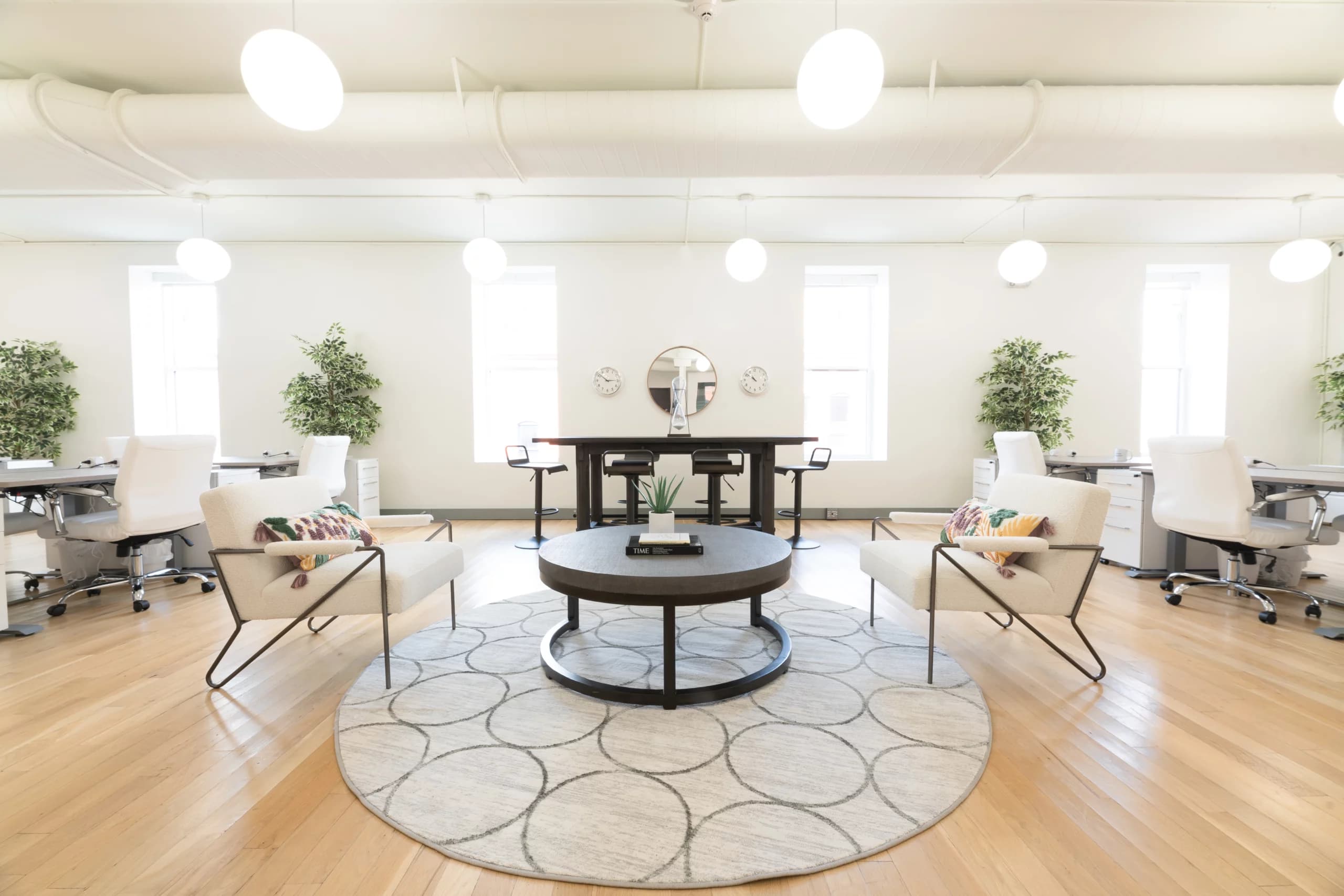 Modern and bright office space featuring a circular area rug, two lounge chairs with a coffee table, surrounded by desks and office chairs, all under soft pendant lighting in a room with white walls and wooden floors.