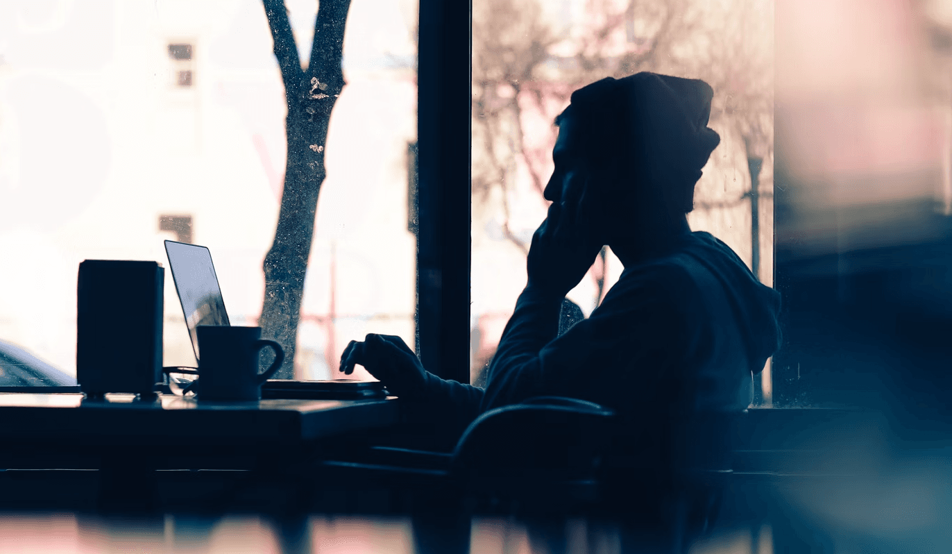 Silhouette of a person wearing a hat, sitting at a cafe table, working on a laptop next to a window with a street view visible in the background.