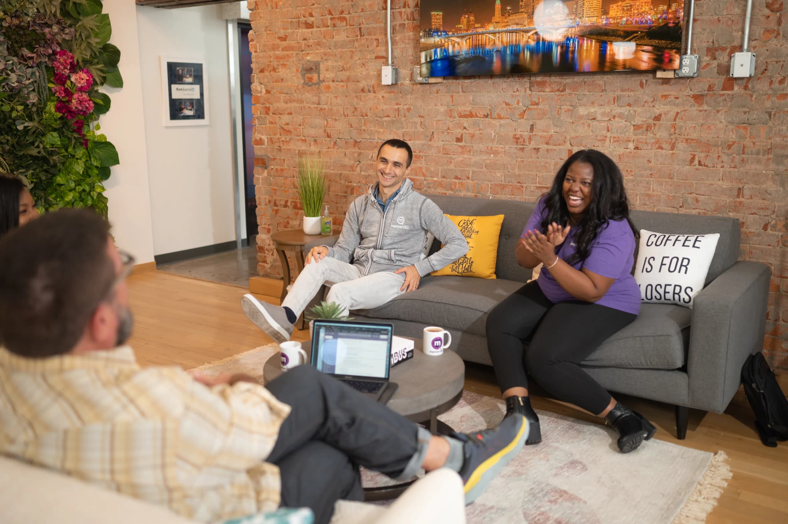 Three colleagues in a casual office lounge with brick walls, engaging in a friendly discussion. One woman is laughing, another man is seated comfortably, and a third is seen from behind, all surrounded by vibrant indoor plants and modern decor.