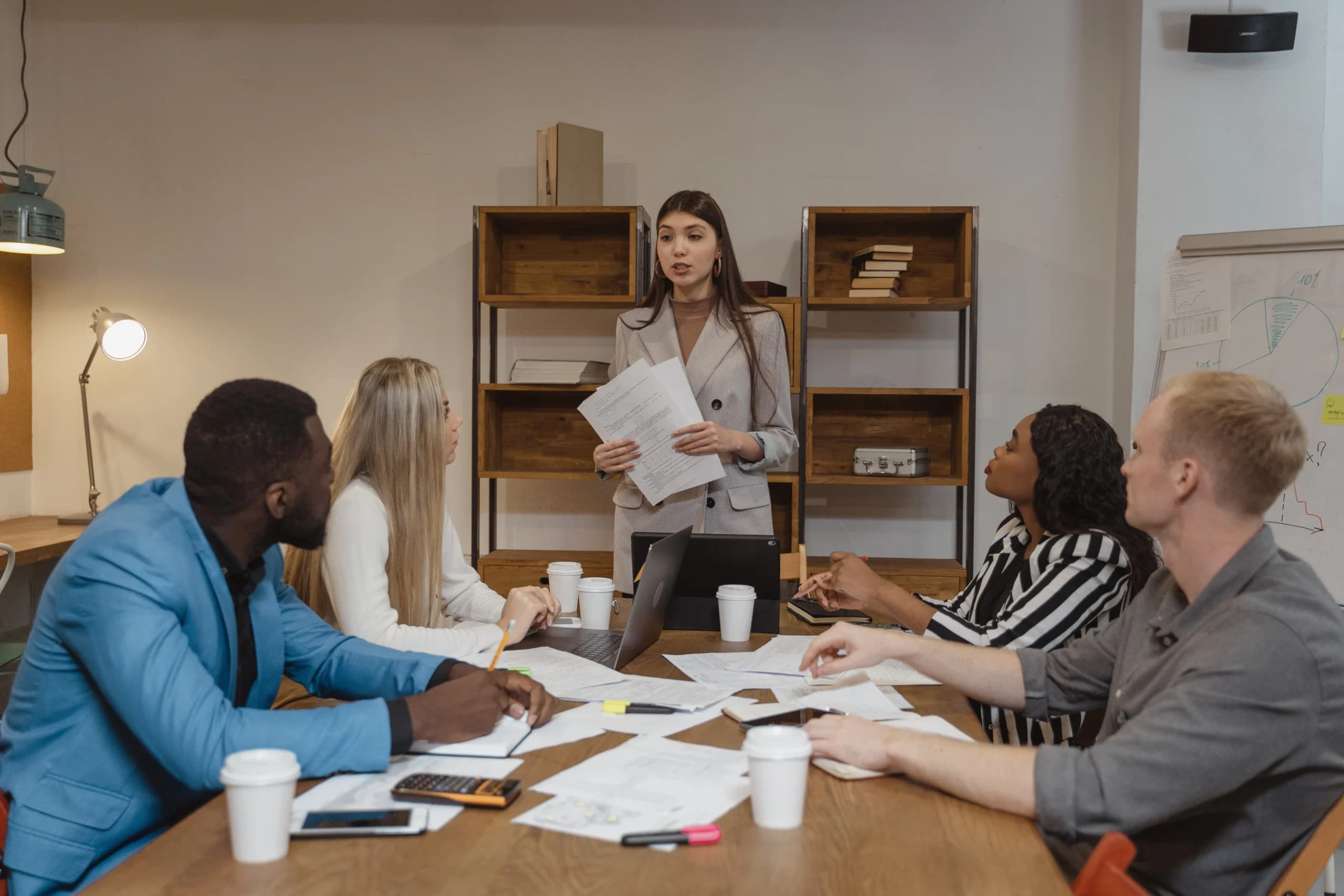 Diverse group of five professionals gathered around a conference table in a well-lit office. A woman stands presenting documents while her attentive colleagues, including men and women of different ethnicities, listen and take notes.