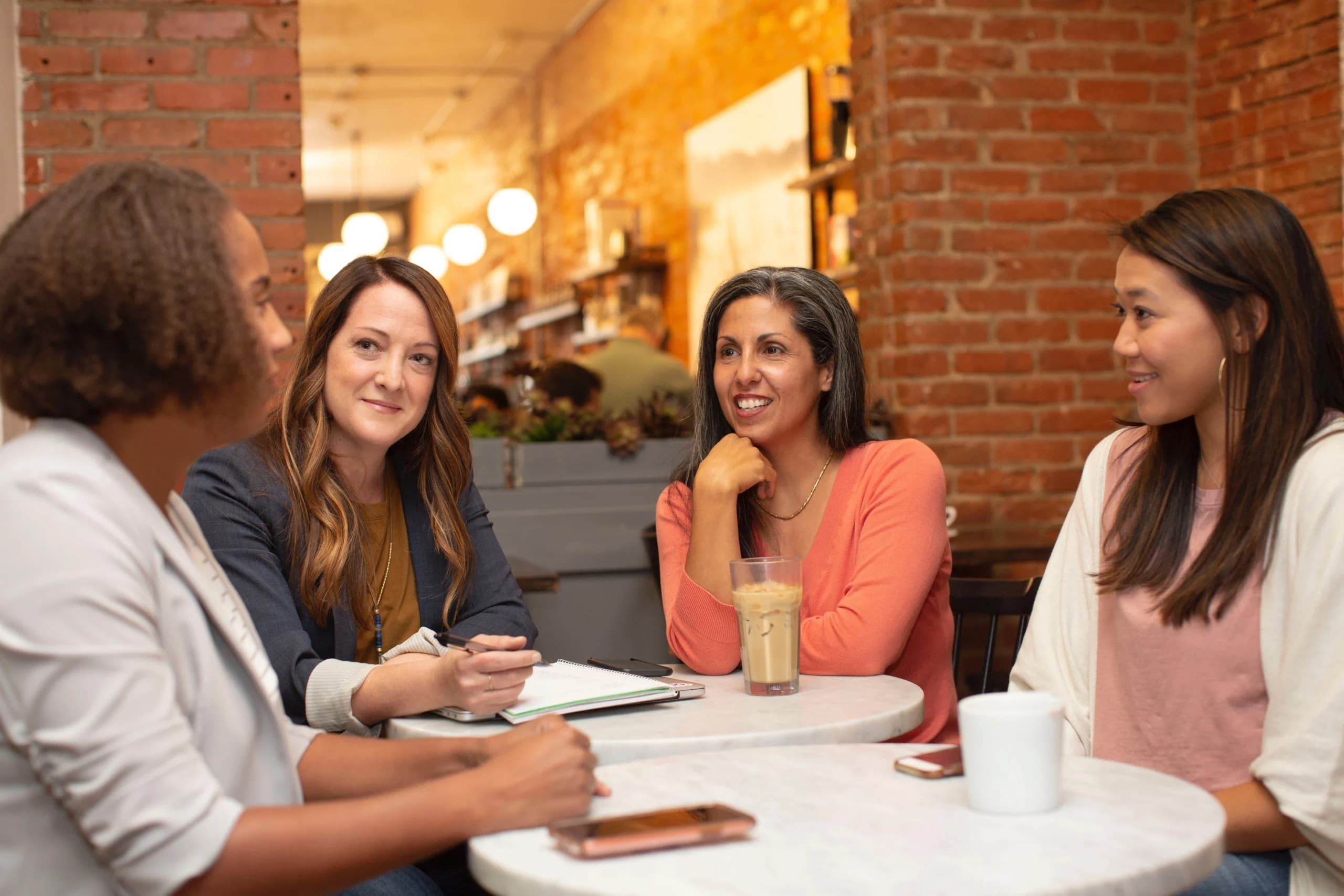 Four diverse women engaging in conversation at a café table, set against a backdrop of a cozy brick-walled interior with ambient lighting.