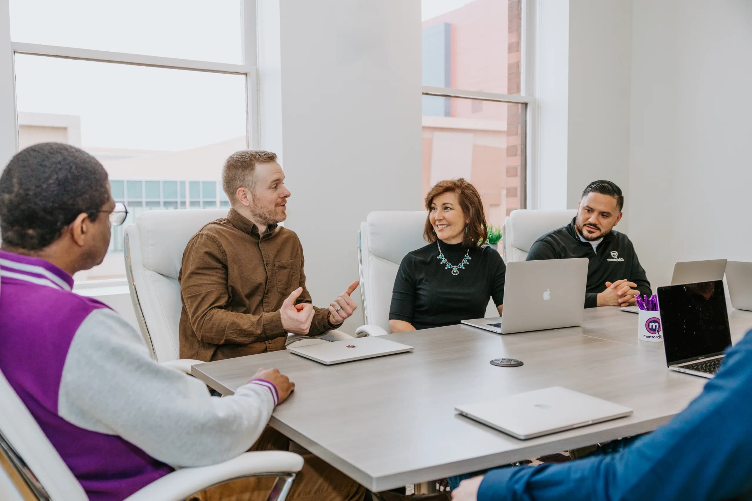 Four professionals in a modern office meeting room, engaging in a discussion around a white table with laptops, under natural light from large windows.