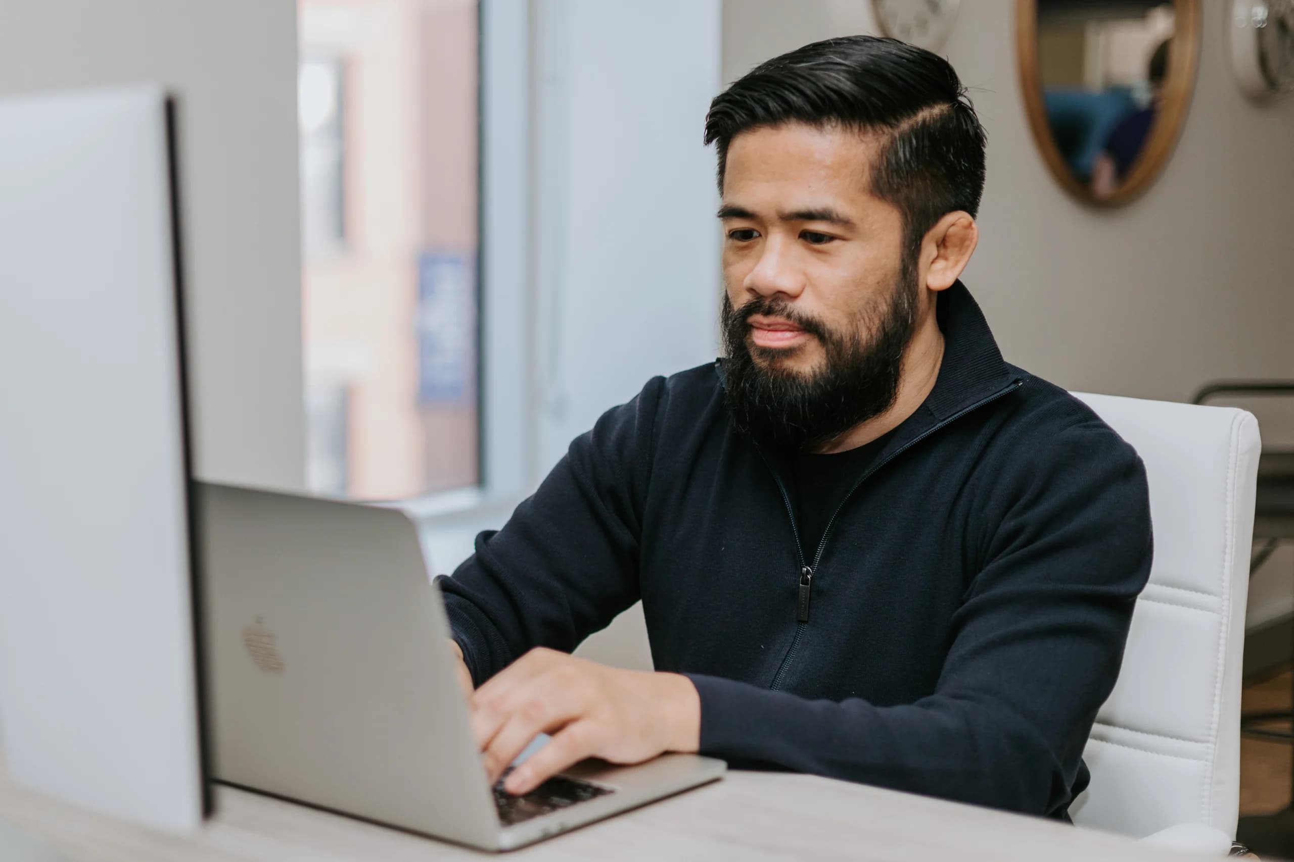 Man with a beard working intently on a laptop at a modern office desk. He wears a dark sweater and the setting includes a bright interior with decorative mirrors in the background.