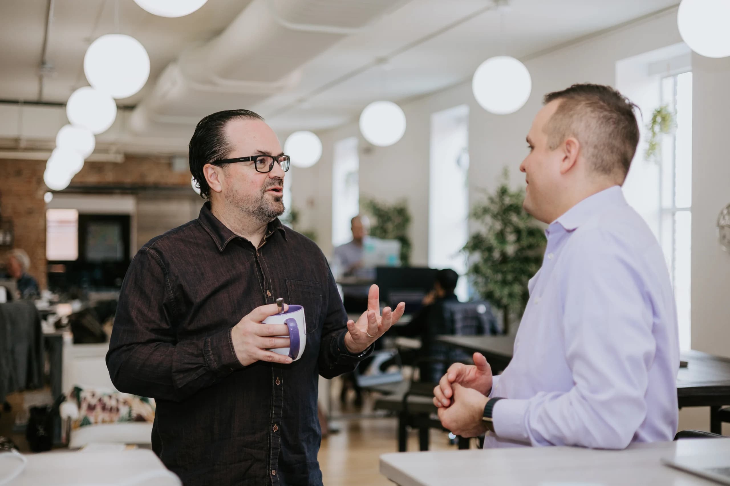 Two men in casual office attire engaging in a discussion in a modern workspace, with one holding a coffee mug. The environment is well-lit with overhead lights and features an open office layout.