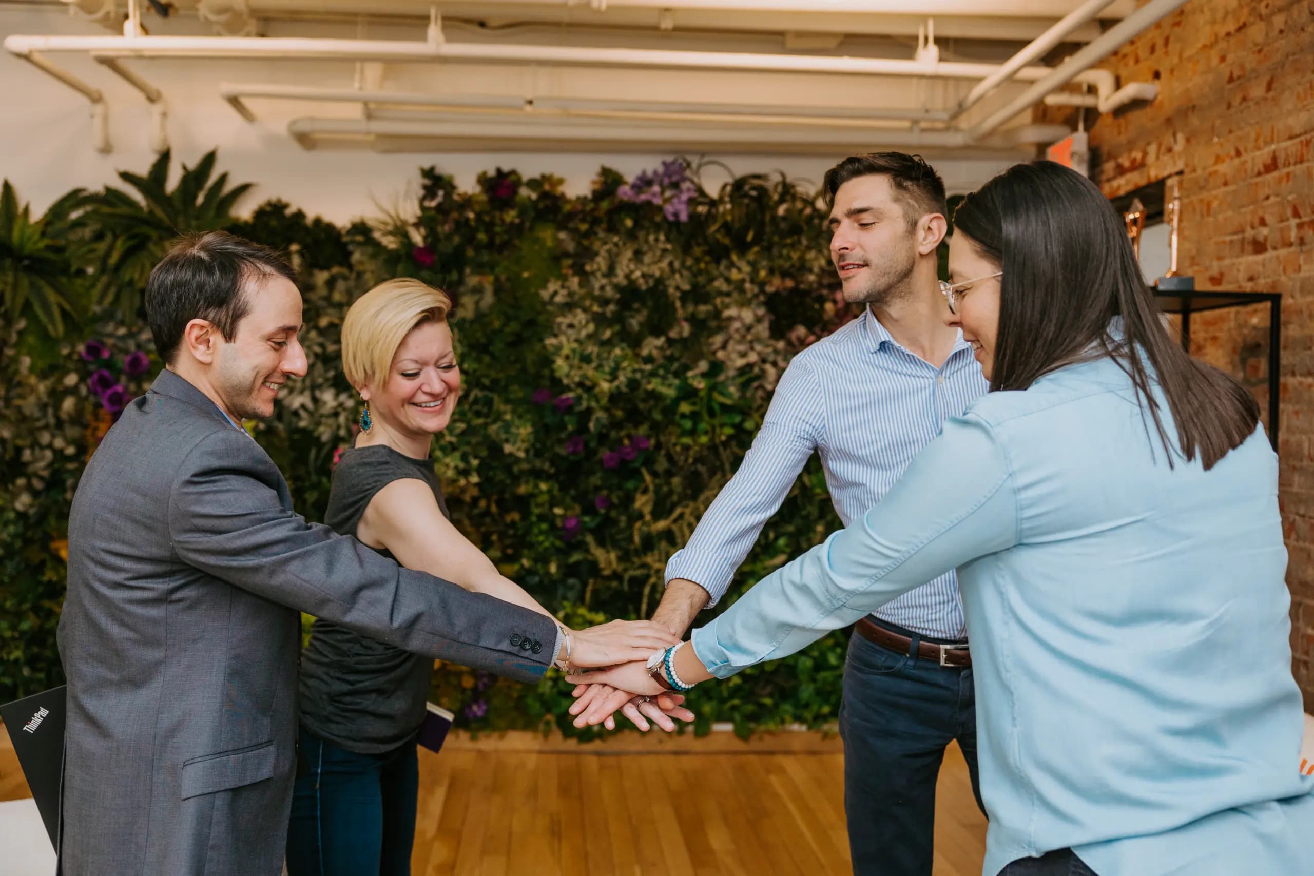 Four colleagues in an office space joyfully performing a team hand stack, surrounded by a lush vertical garden. They appear cheerful, showcasing a sense of teamwork and unity.