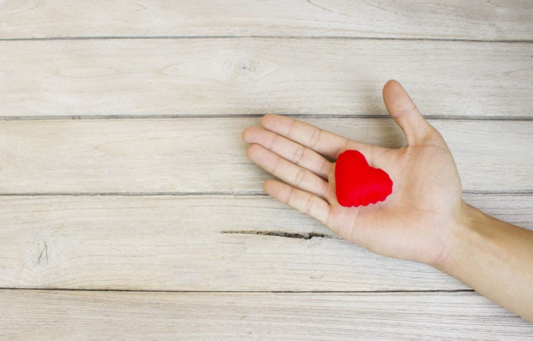 A human hand holding a small red stuffed shaped heart in their palm against a wood plank background.