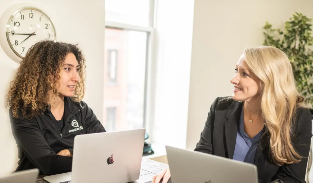 Two female employees chat in an office with computers open. 