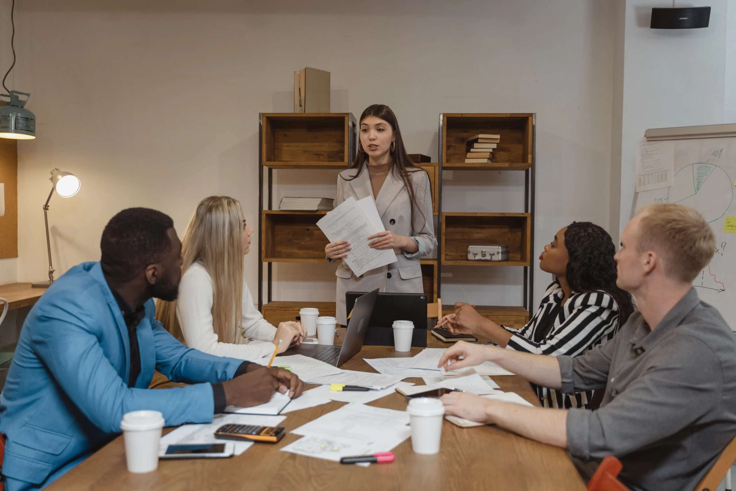 In a meeting room, a team listens to a standing woman presenting leadership 101 papers. The table is scattered with notes and laptops; a whiteboard with graphs is in the background, indicating an ongoing work discussion.