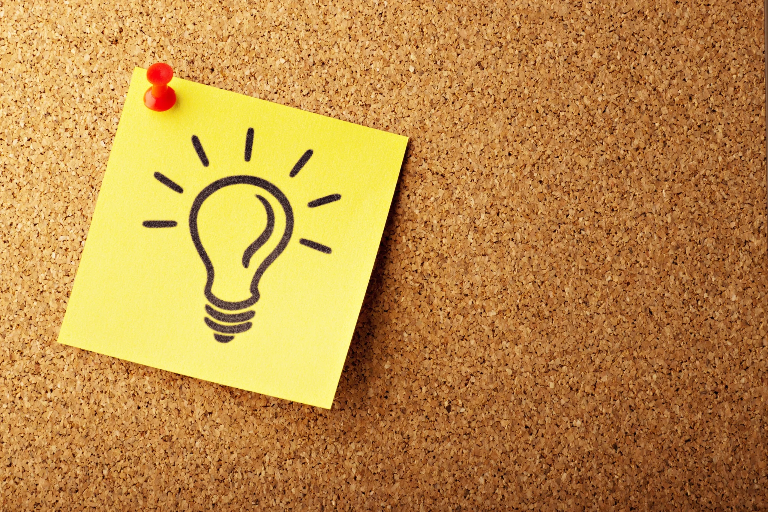 A yellow sticky note with a hand-drawn light bulb symbol is pinned to a cork board, symbolizing an idea or inspiration.