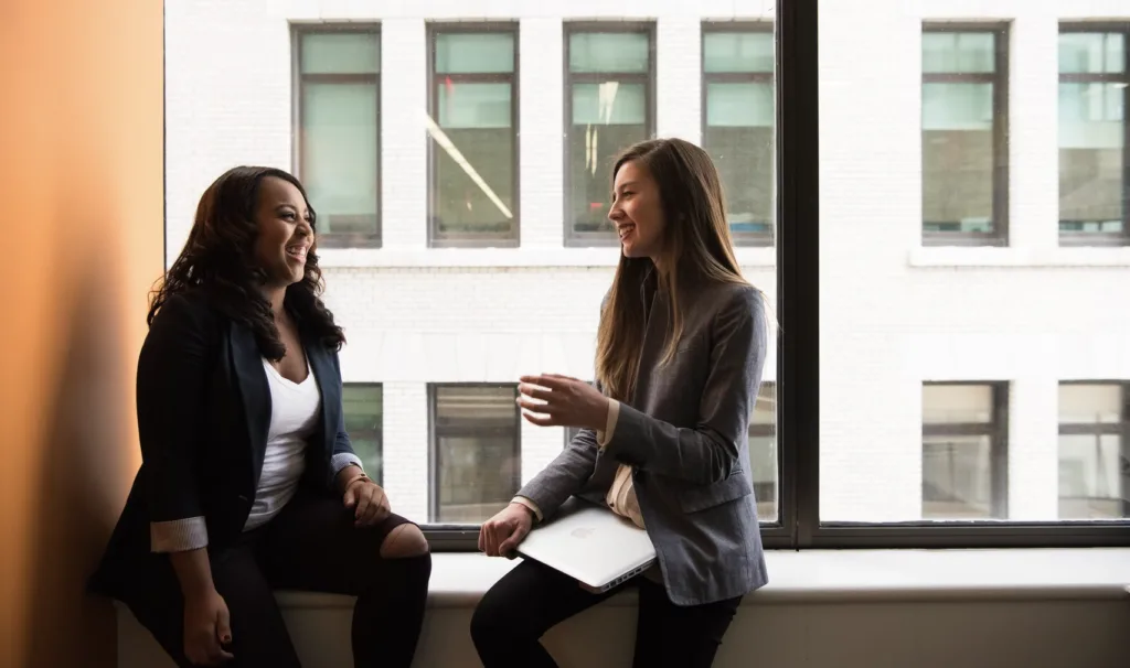 An image of two women sitting in a window sill in an office. They are discussing important topics, such as mentoring vs coaching, while also laughing together. 
