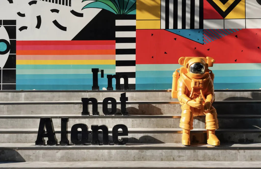 Artistic image of an orange astronaut sitting on concrete steps sitting by a sign that says "I'm Not Alone"