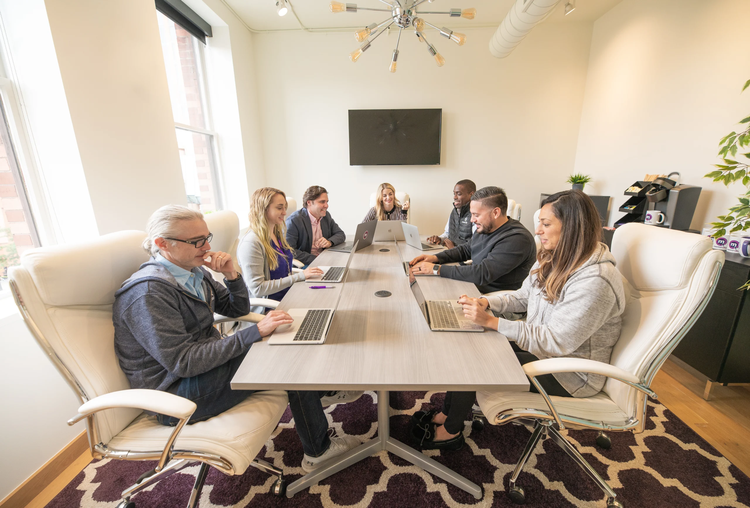 Group of seven diverse professionals in a bright meeting room, sitting around a long table, each working on their laptop, appearing engaged in a collaborative work environment.