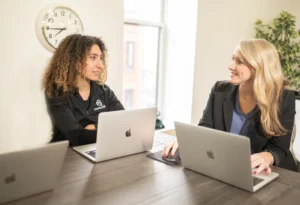 What Is Succession Planning? Key Definition and Strategies 5 Two women working on laptops across from each other at an office table, engaged in a conversation. One woman has curly hair and wears a black shirt, while the other, with blonde hair, wears a business suit.