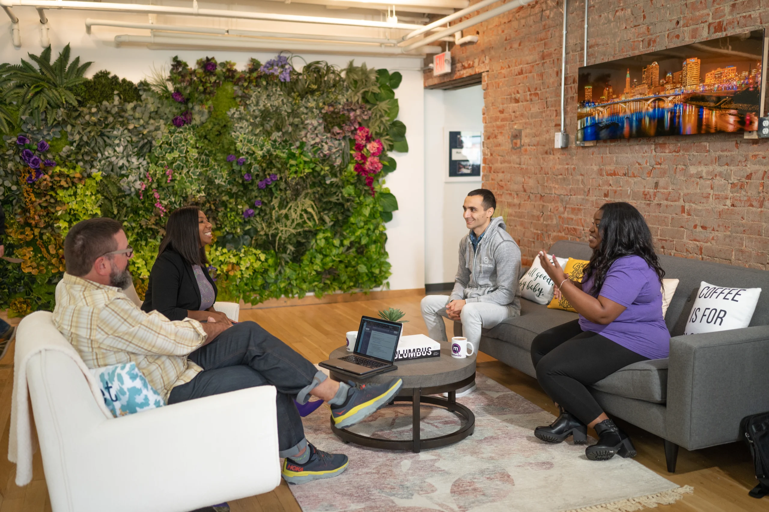 A group of four people in a casual office meeting space. One wall is covered with a lush living plant installation, adding vibrancy to the room. Two men and two women are engaged in a relaxed conversation, with one man sitting on a sofa and holding a cushion with the phrase 'Coffee is for closers'. There's a laptop on a central round table, and the room has a homely feel with its exposed brickwork and a cityscape artwork on the wall.