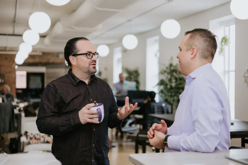 Performance Coaching at Work: What, Why, and How to Do It in the Workplace 5 Performance coaching meeting between two men standing in an office chatting.