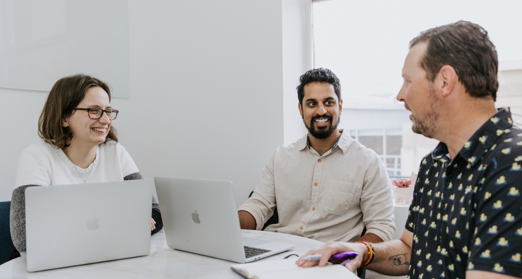 Three colleagues, two men and one woman, smiling and discussing work at a white table with laptops in a bright office setting.