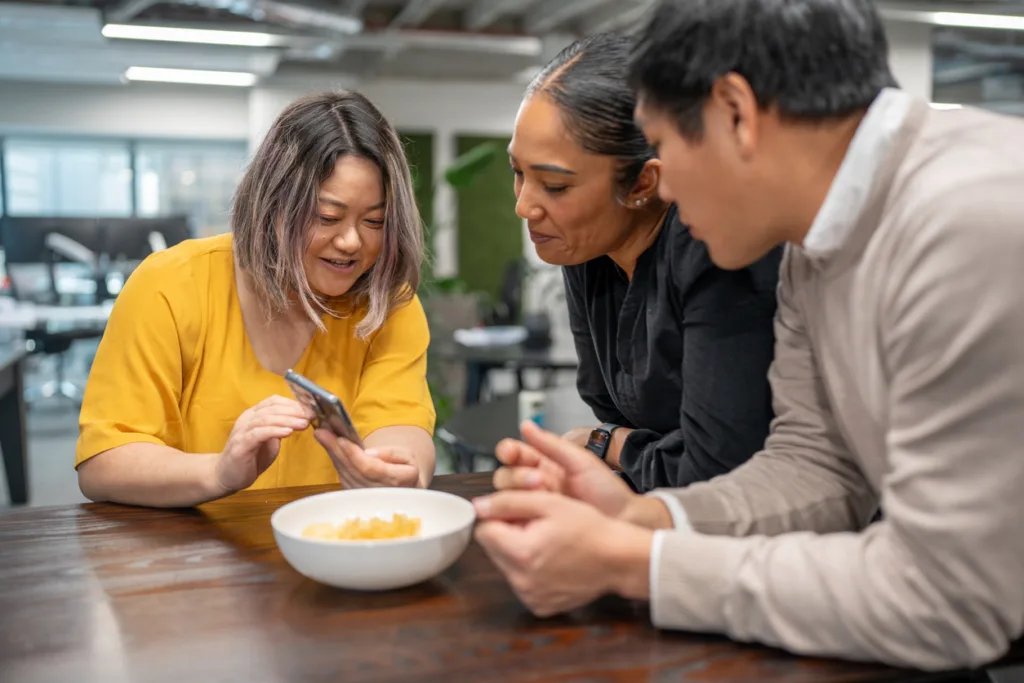 A MentorcliQ Lock Picking Experience 4 Three coworkers gather around a table in an office kitchen, looking at a smartphone together after an event about lock picking